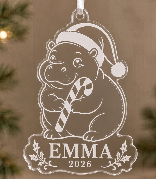 Clear acrylic Santa hippo Christmas ornament laser engraved with a baby hippo in a Santa hat holding a candy cane, holly sprigs, and a name banner reading EMMA 2026, hanging from a white ribbon